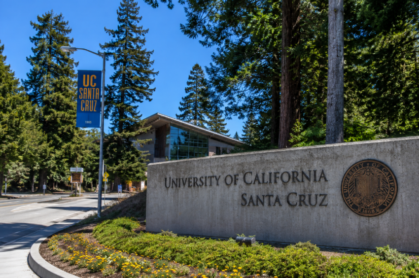 Entrance to the University of California, Santa Cruz campus with the university sign, redwood trees, and a modern campus building.