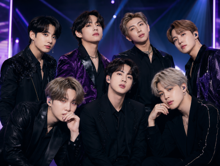 Seven members of a K-pop boy band posed together in stylish dark stage outfits under purple and blue concert lighting.