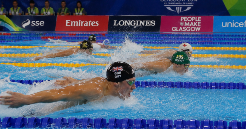 Swimmers compete in a Commonwealth Games race in Glasgow as athletes power through the pool beneath event banners and crowded spectator stands.