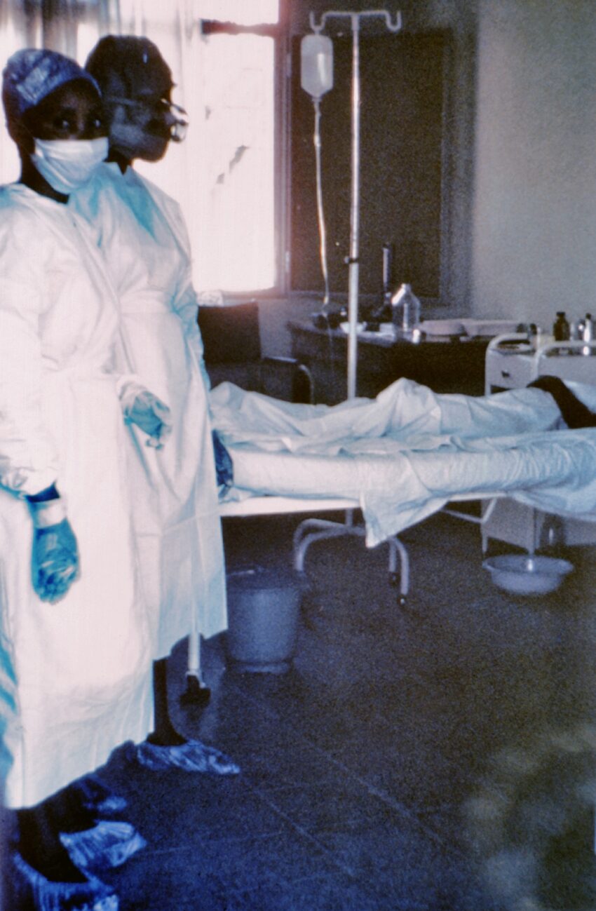 This 1976 photograph shows two nurses standing in front of Ebola case #3, who was treated, and later died at Ngaliema Hospital, in Kinshasa, Zaire. Ebola hemorrhagic fever (Ebola HF) is a severe, often-fatal disease in humans, chimpanzees, gorillas and orangutans that has appeared sporadically since its initial recognition in 1976.