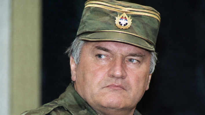 Documentary-style portrait of Ratko Mladic in camouflage military uniform and cap, shown with a stern expression against a dark blurred background.