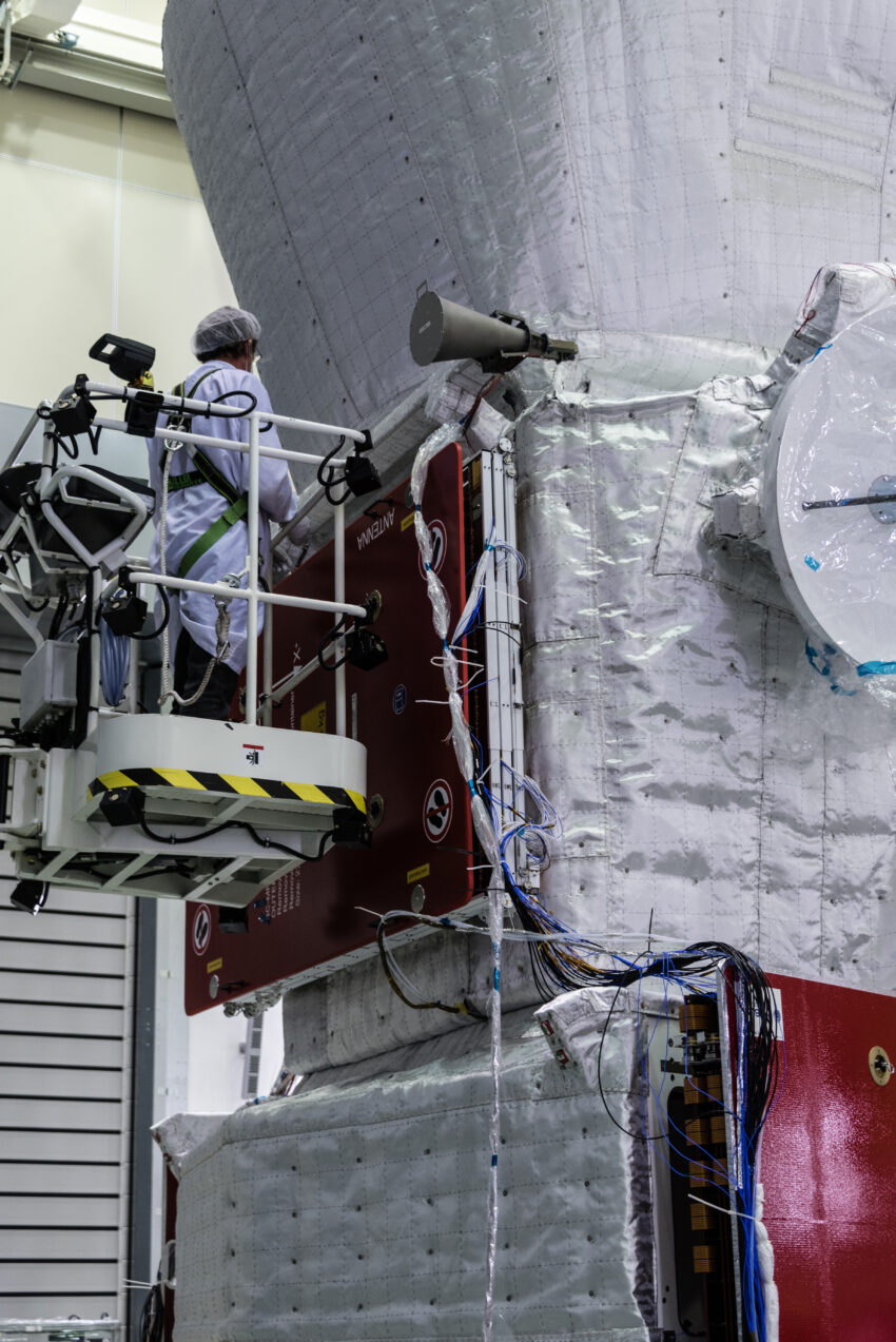 Examining the BepiColombo stack in ESA’s test centre. The person gives a sense of scale of the full spacecraft stack, which is more than 6 m high. Although not fully visible in this view, the Mercury Transfer Module is at the bottom, the Mercury Planetary Orbiter is in the middle, and the Mercury Magnetospheric Orbiter’s Sunshield and Interface Structure is seen at the top. The red panels are covers protecting the folded solar wings, and will be removed before flight. The white covering on the spacecraft is a thermal blanket to help protect the craft from the high temperatures that will be experienced operating close to the Sun. The horn-shaped object on the corner of the Mercury Planetary Orbiter is the spacecraft’s medium gain antenna; it is fixed to a boom that will be deployed after launch.