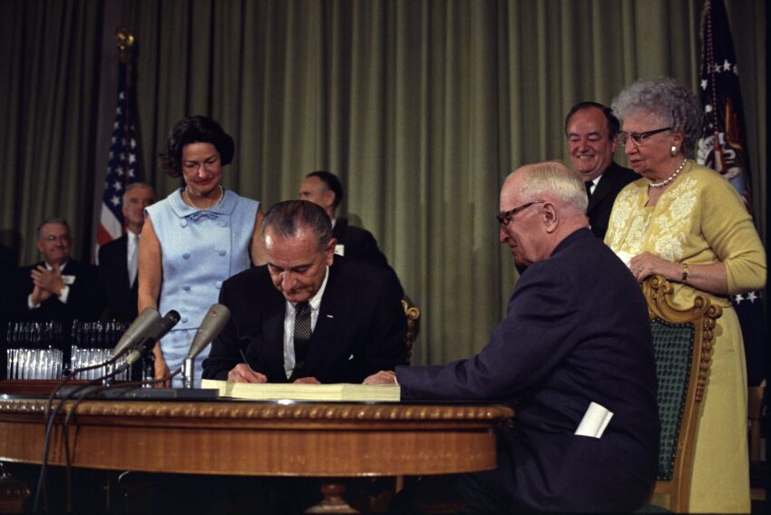 President Lyndon B. Johnson signing the Medicare Bill at the Harry S. Truman Library in Independence, Missouri. Former president Harry S. Truman is seated at the table with President Johnson. The following are in the background (from left to right): Senator Edward V. Long, an unidentified man, Lady Bird Johnson, Senator Mike Mansfield, Vice President Hubert Humphrey, and Bess Truman.